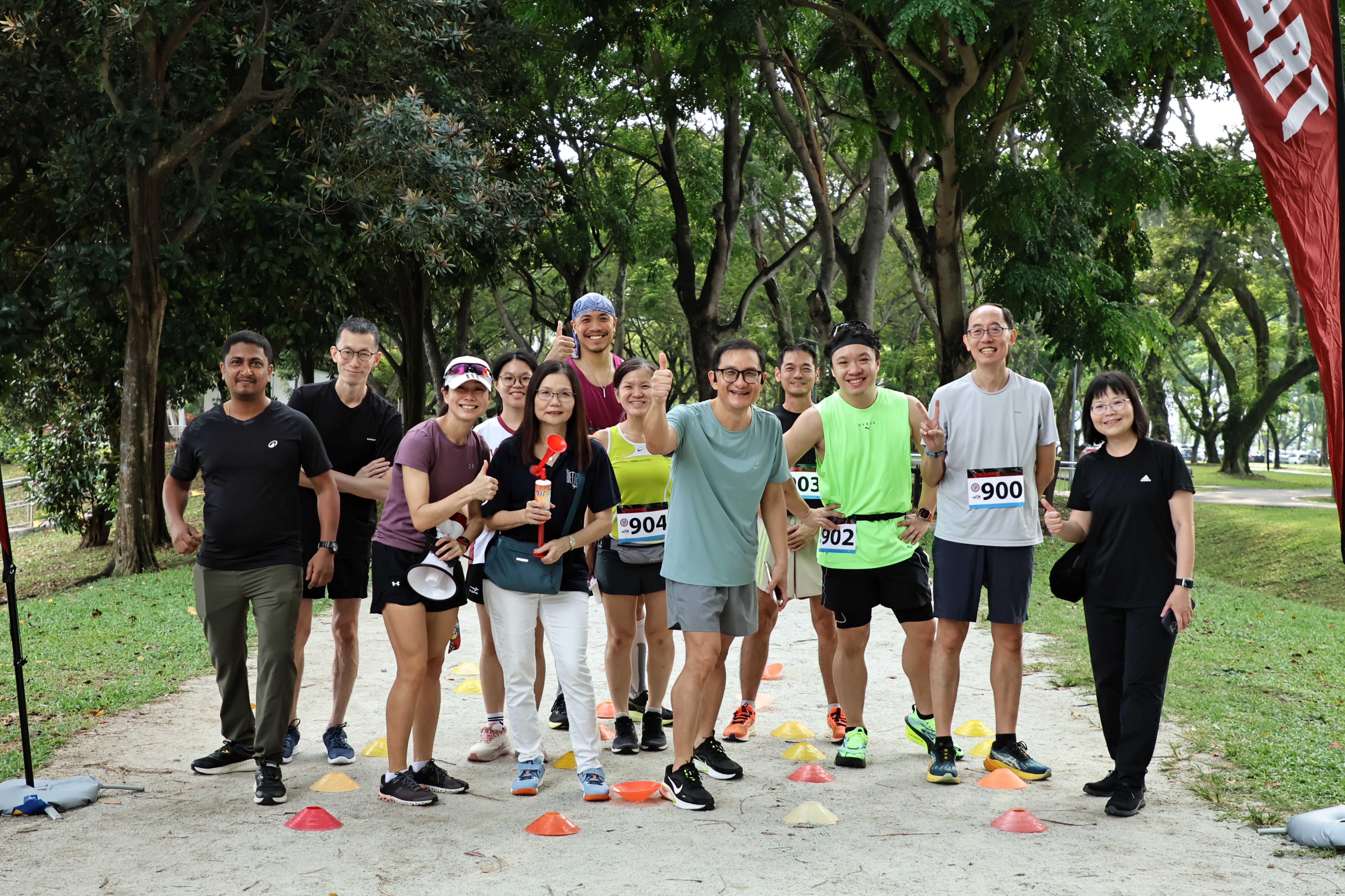 A group of about 12 TMJC staff members pose together outdoors under shady trees, some wearing race bibs, smiling and giving thumbs up before a run.